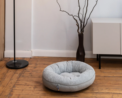 Round gray pet bed on a wooden floor with a vase and plant in the background.